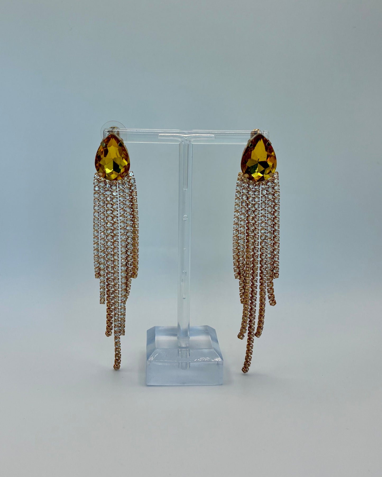 Pair of earrings with yellow gemstones and chain tassels on a clear stand against a light gray background.