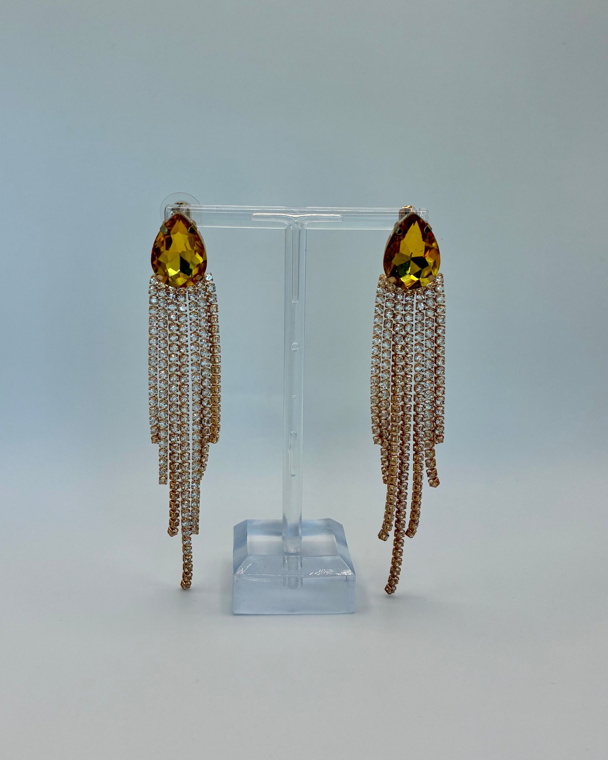 Pair of earrings with yellow gemstones and chain tassels on a clear stand against a light gray background.
