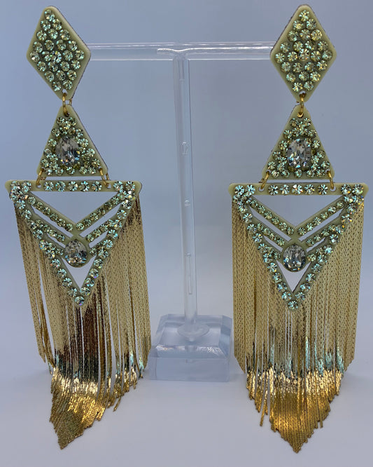Gold fringe earrings with green gemstones on a clear stand against a gray background