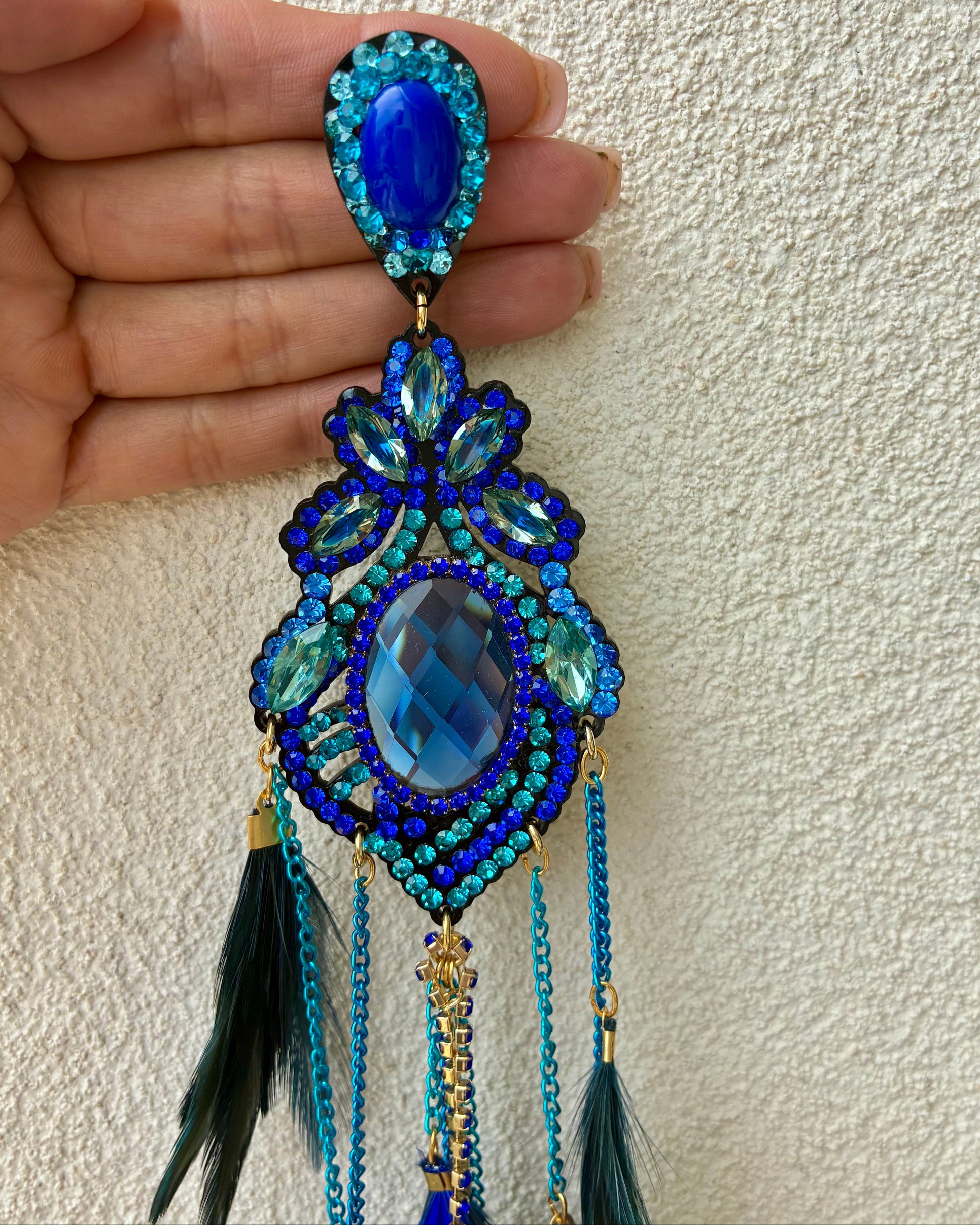 Blue gemstone earring with feather details held by a hand against a beige background