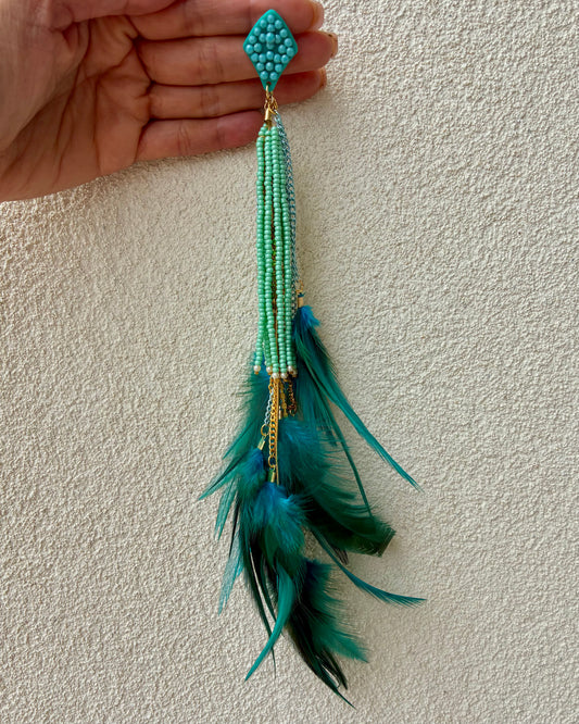 Turquoise beaded earring with feathers held against a beige wall.