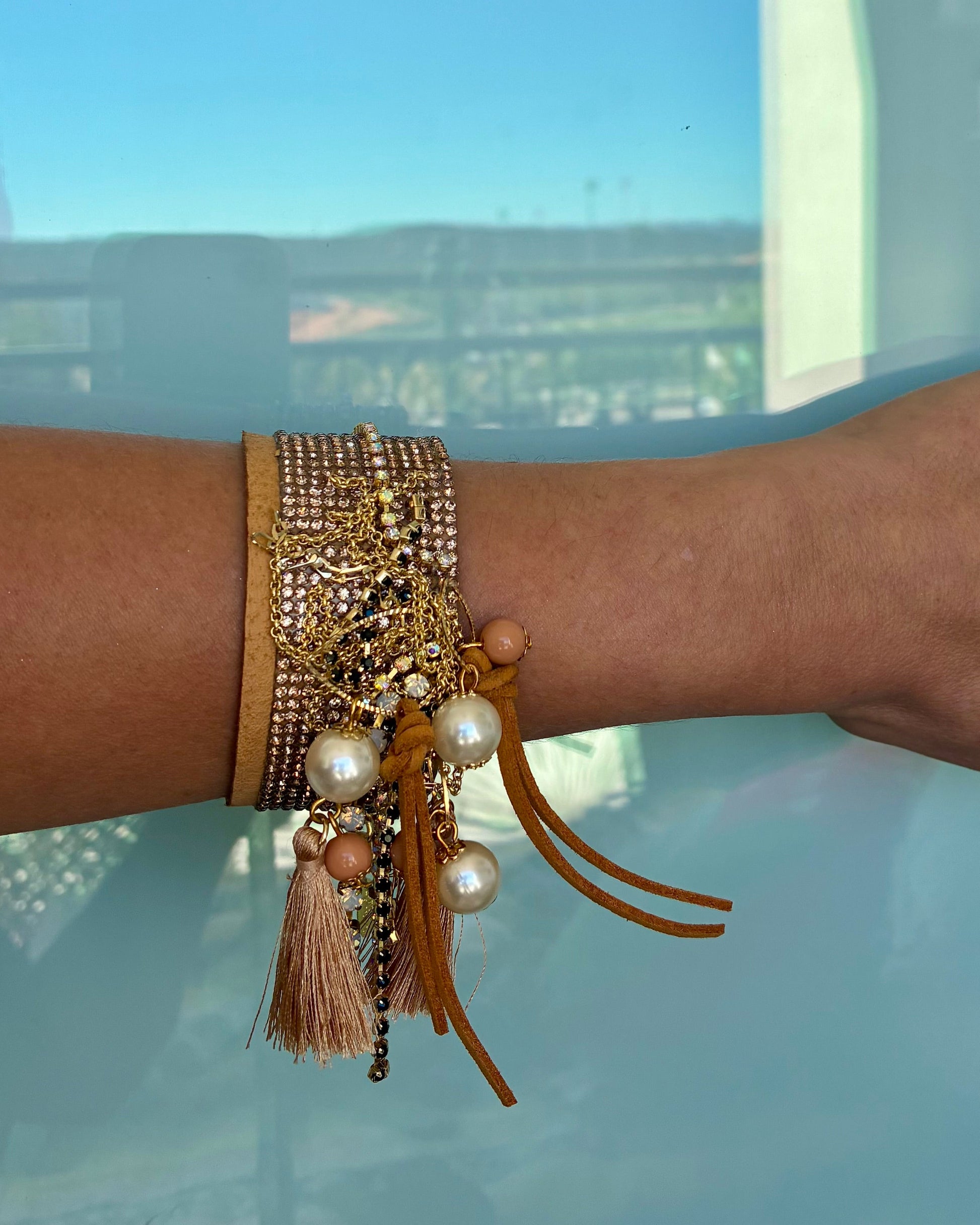 Close-up of a wrist wearing multiple gold and pearl bracelets with tassels against a blurred outdoor background.
